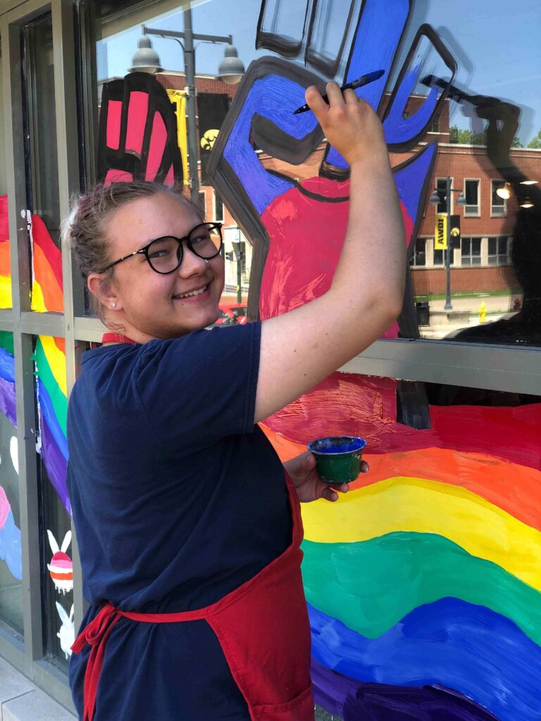 A photo of Phil (a white person with glasses and light brown hair up in a bun) depicts him wearing a dark blue shirt and a red apron. He is painting a fist emerging from a rainbow. 